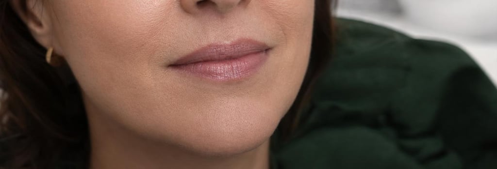 Photorealistic close-up of lips in soft clinical lighting: mature lips showing natural skin texture, subtle smile, no exaggerated volume, shallow depth of field, warm neutral tones, forest green fabric in background, editorial clinical-luxury feel, no branding, no text.