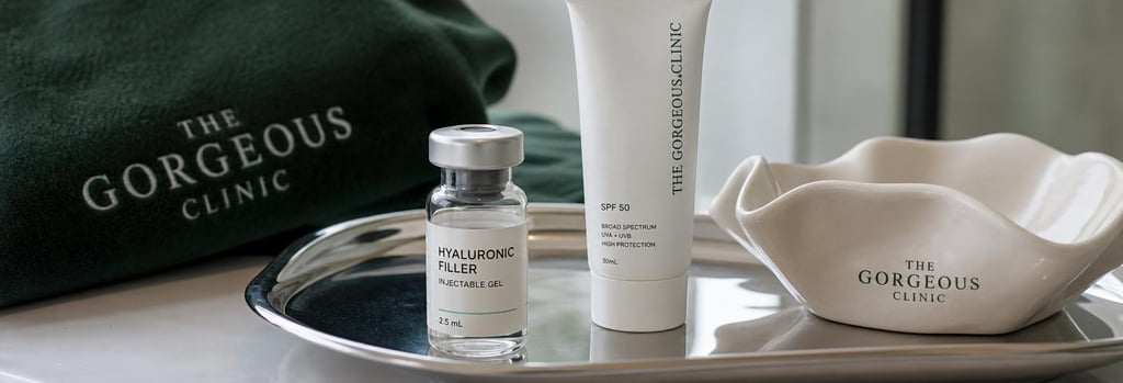 Photorealistic close-up of a glass vial of hyaluronic filler on a polished tray, alongside a small SPF tube and folded cream ceramic dish, soft natural clinic light, forest green fabric in background, editorial composition.