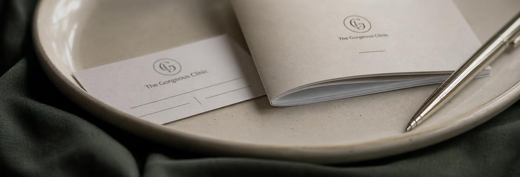 Photorealistic close-up of an appointment card, a folded consent form and a pen on a cream ceramic tray, subtle forest green cloth beneath, soft editorial lighting, single focal point on the consent form edge, no readable text.